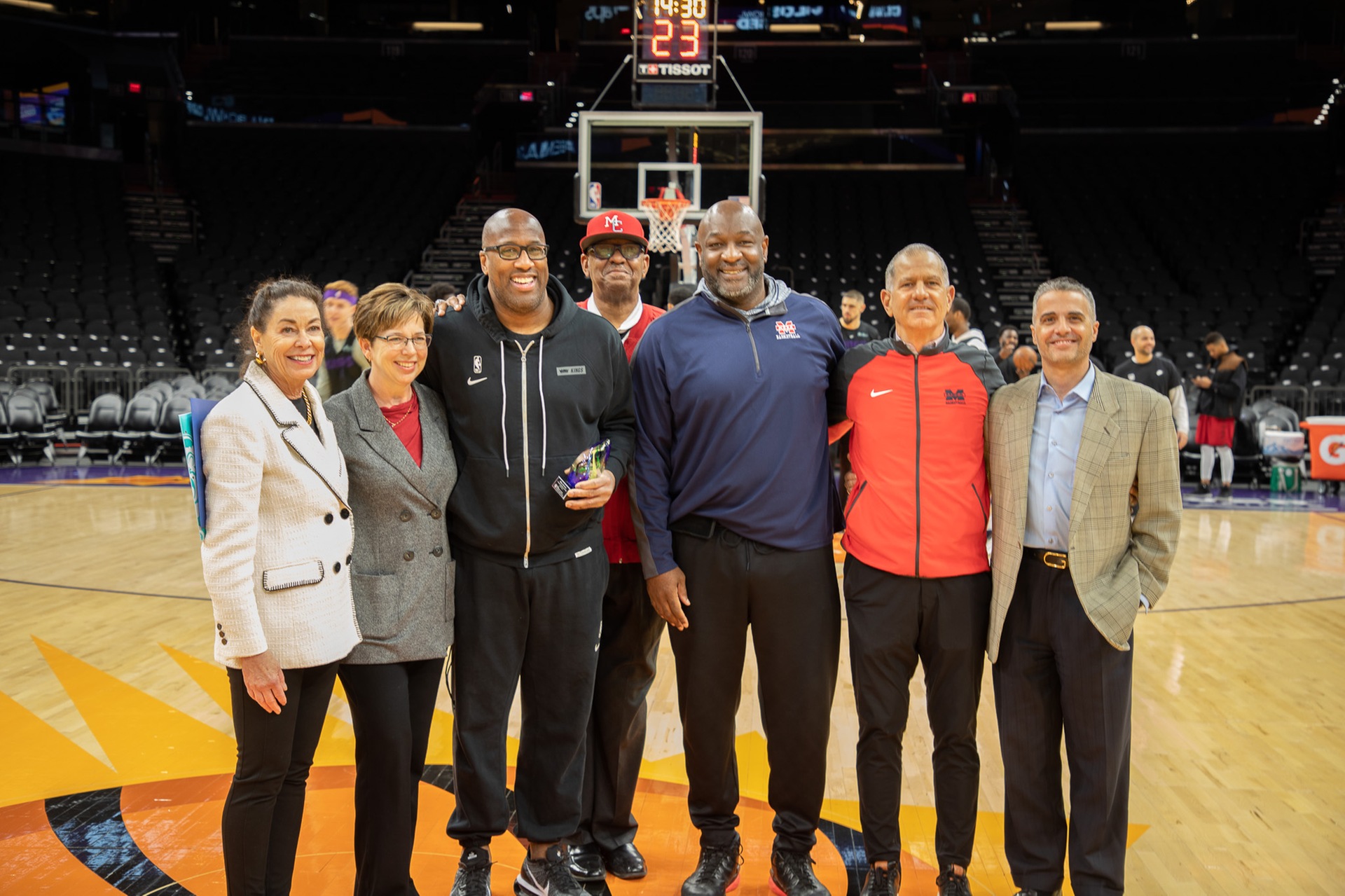 Group photo on a professional basketball court featuring seven individuals standing together near center court beneath a basketball hoop and shot clock. The group poses for a recognition moment before a game, with several wearing Mesa Community College apparel. Arena seating and players warming up are visible in the background. One individual in the center holds a commemorative plaque while the group smiles for the photo.
