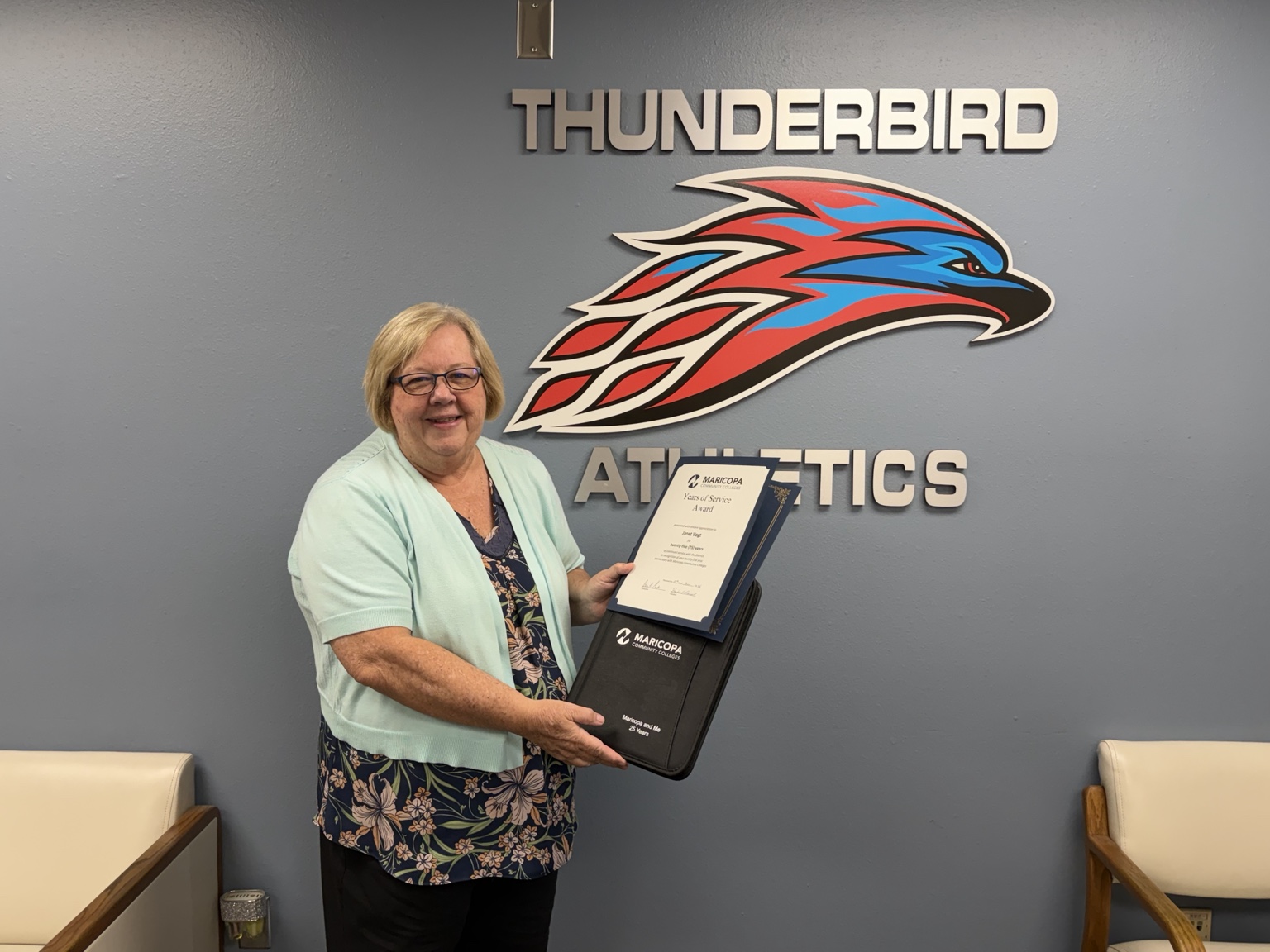  Administrative Secretary Janet Vogt smiles while holding her 25 Years of Service Award and commemorative gift in front of the Thunderbird Athletics wall at Mesa Community College. The large Thunderbird logo and 
