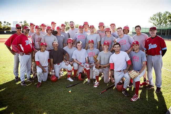  Group photo of the Mesa Community College baseball team and coaching staff standing and kneeling on the field during practice. Players wear gray practice shirts, red caps, and baseball pants while coaches stand at either end of the group in red tops. The photo was taken during the filming of Red Bull's viral 