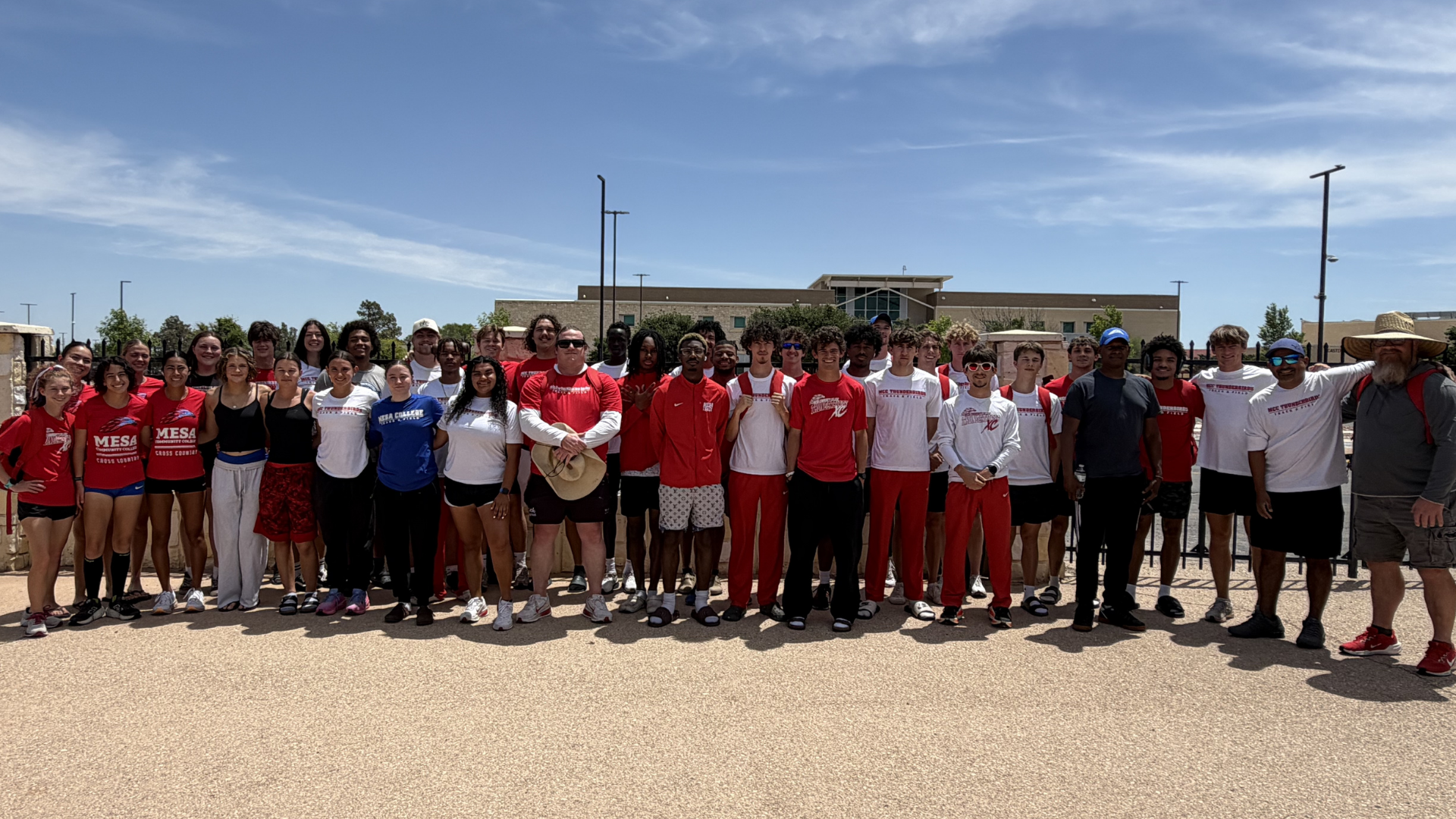 Members of the Mesa Community College cross country and track & field programs pose together outdoors on campus under a bright blue sky. Student-athletes and coaches wear a mix of Mesa Thunderbirds apparel in red, white, and blue while standing in front of a campus building during a team gathering.

